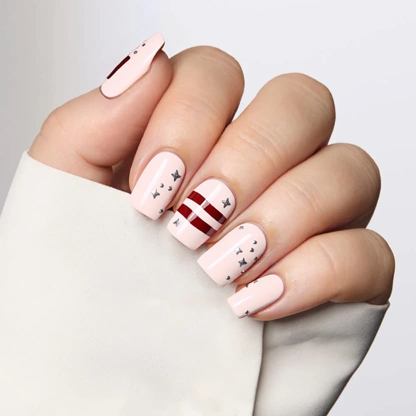 Nails adorned in a soft pastel pink base, serving as a warm canvas that radiates elegance. Two accent nails feature alternating dainty stripes and playful spots in a muted red, maintaining a whimsical yet refined aesthetic. Delicate silver stars are sprinkled across the design, adding a touch of sparkle without overwhelming the overall look. The other nails remain in a seamless pink finish, creating a cohesive and visually balanced composition that complements the graceful curves of medium almond nails.