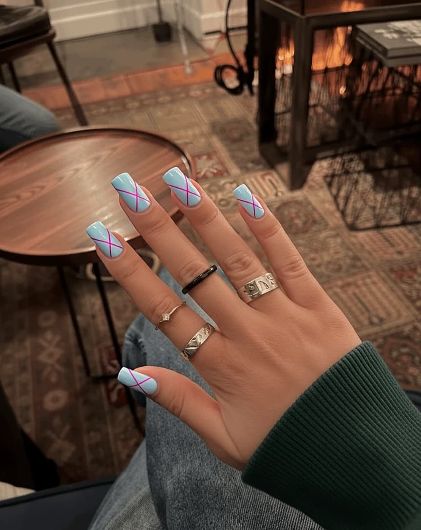 Nails featuring a soft pastel palette with a base color of light blue across most nails, complemented by delicate geometric patterns in pink and lilac. The index and ring fingers showcase a minimalist design with intersecting triangles and thin lines creating a modern, abstract look, while the other nails remain a smooth, solid light blue. The geometric elements are kept subtle and sparse, with no more than four shapes per accented nail, ensuring an elegant and feminine finish. A glossy top coat enhances the overall appearance, adding a fresh and airy vibe to these medium-length square nails.
