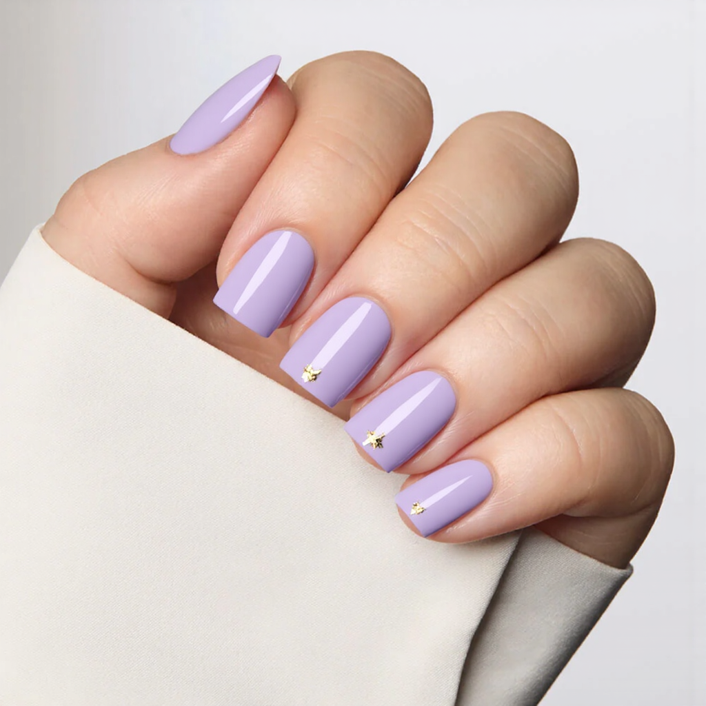 Short square nails in a pastel purple, each with a tiny gold foil accent on one corner.