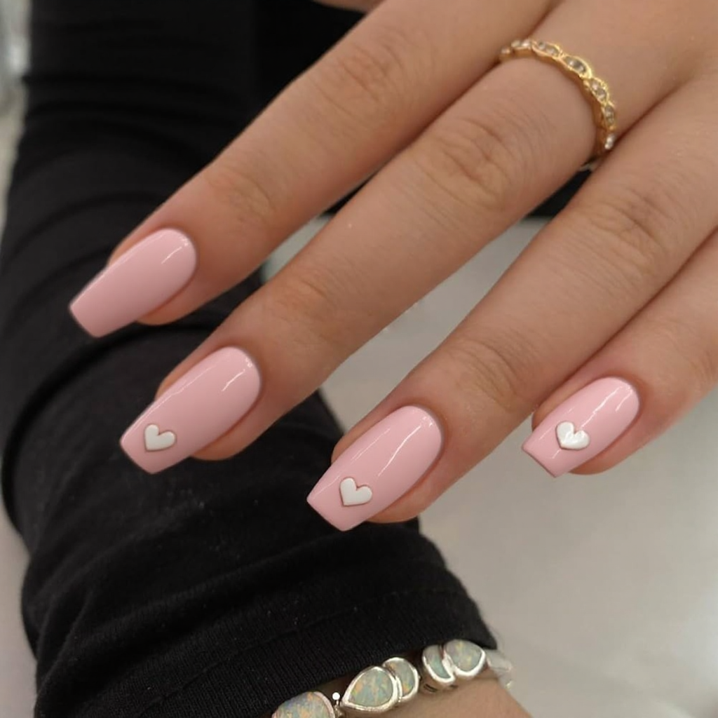 Short stiletto nails in baby pink, each featuring a small heart decal near the tip for extra cuteness.