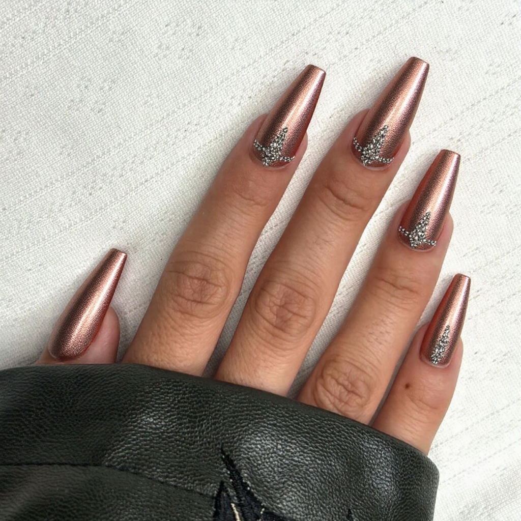 Long stiletto nails in a metallic rose gold, each ring finger featuring a line of rhinestones down the center.