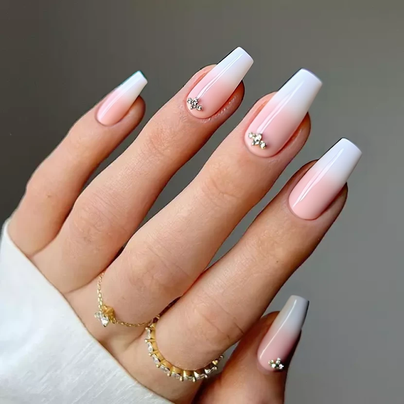 Nails painted in a soft pastel pink base that exudes elegance, with a glossy finish enhancing the delicate hue. On the middle fingers, minimalist designs feature a subtle gradient shifting into a gentle blush, adorned with a few small, sparkling rhinestones clustered near the cuticle, creating a sophisticated look. The other nails are left in a pristine pastel pink to maintain a harmonious balance, perfect for medium stiletto nails.