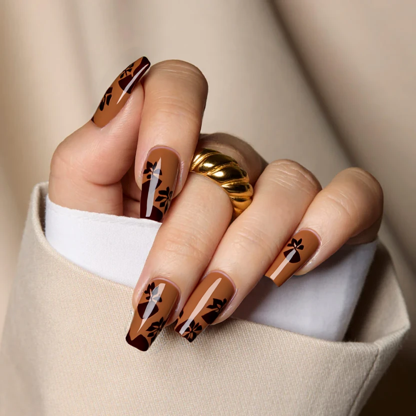 Nails adorned in a soft, muted caramel brown as a base color, creating an elegant and minimalistic look perfect for autumn. Three nails on each hand maintain the warm caramel hue, while an accent of delicate leaf patterns in a deeper chocolate brown is featured on the ring fingers, with no more than four small leaves gracefully scattered. The overall design is unified with a glossy finish, enhancing the sophisticated and seasonal aesthetic while beautifully complementing the structured lines of medium square nails.