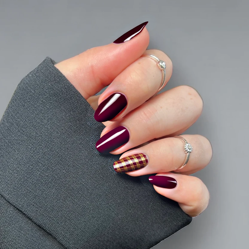 All nails are elegantly coated in a rich, glossy burgundy polish, enhancing the sophisticated allure of the almond shape. On one hand, the ring finger features a chic accent nail adorned with a minimal plaid design, created by thin gold lines delicately crossing over the burgundy base. This festive pattern maintains a refined balance, allowing the other nails—solid burgundy in color—to provide a simple yet classy backdrop. The combination of the deep burgundy and the subtle gold plaid ensures that the design is both striking and effortlessly elegant, perfectly suited for the short almond nails.