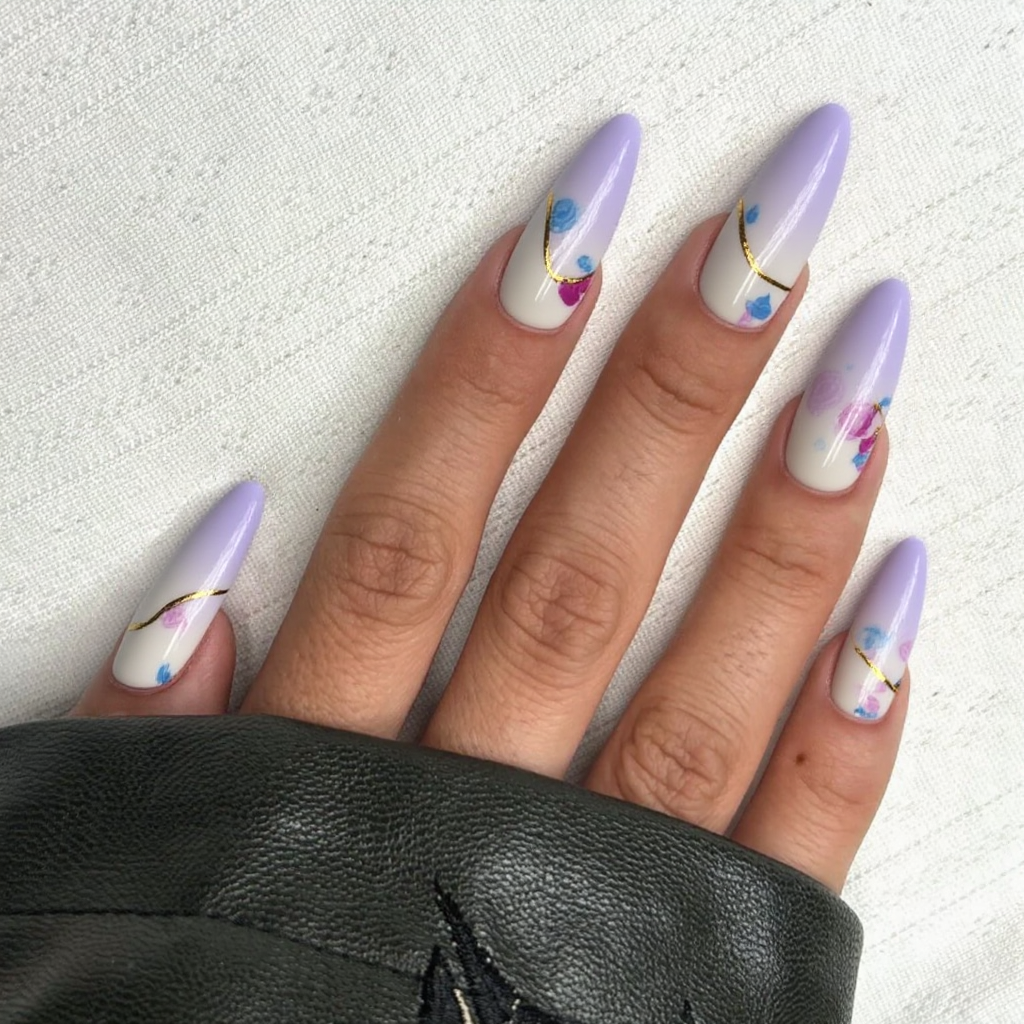 Long stiletto nails featuring a delicate ombre effect that transitions from soft lavender to creamy white, creating a dreamy and ethereal base. On the index fingers, a subtle metallic gold line gently arcs from the base to the tip, adding an element of elegance without overwhelming the design. The remaining nails are adorned with minimalistic pastel-colored abstract shapes in muted pink and blue, scattered sparsely for a balanced, artistic touch. The finish is glossy, enhancing the overall sophisticated vibe while ensuring the design remains unforgettable yet feminine.