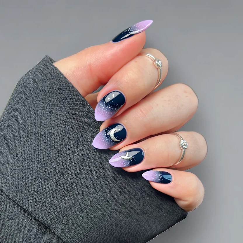 Nails featuring a dark blue base that transitions into a soft purple gradient towards the tips, capturing the essence of a cosmic night sky. On the thumb and ring finger, delicate speckles of fine silver glitter are applied to create a starry effect, twinkling softly like distant celestial bodies. A petite hand-painted crescent moon in silver accents the ring finger, adding a whimsical touch, while the remaining nails maintain a smooth gradient finish with sparkling elements. The soft almond shape enhances the overall elegance, providing a serene yet enchanting cosmic vibe in a practical design suited for short nails.