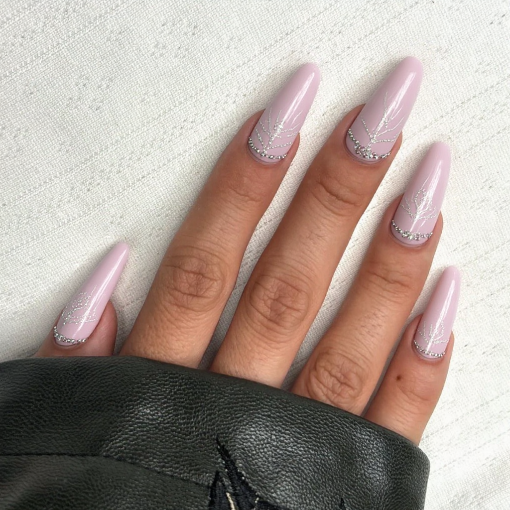 Stiletto nails adorned in a soft pastel pink base, lending an elegant backdrop for the artistic design. On the middle and ring fingers, a minimalist geometric pattern incorporates delicate lines, forming a subtle asymmetrical design in a pearlescent white that subtly catches the light. Tiny, iridescent rhinestones are strategically placed along the edges of the design for added sophistication without overwhelming the overall look. The other nails are kept plain pastel pink, maintaining a harmonious balance. A glossy finish enhances the elegance, making this design both feminine and memorable while perfectly highlighting the dramatic shape and length of long stiletto nails.