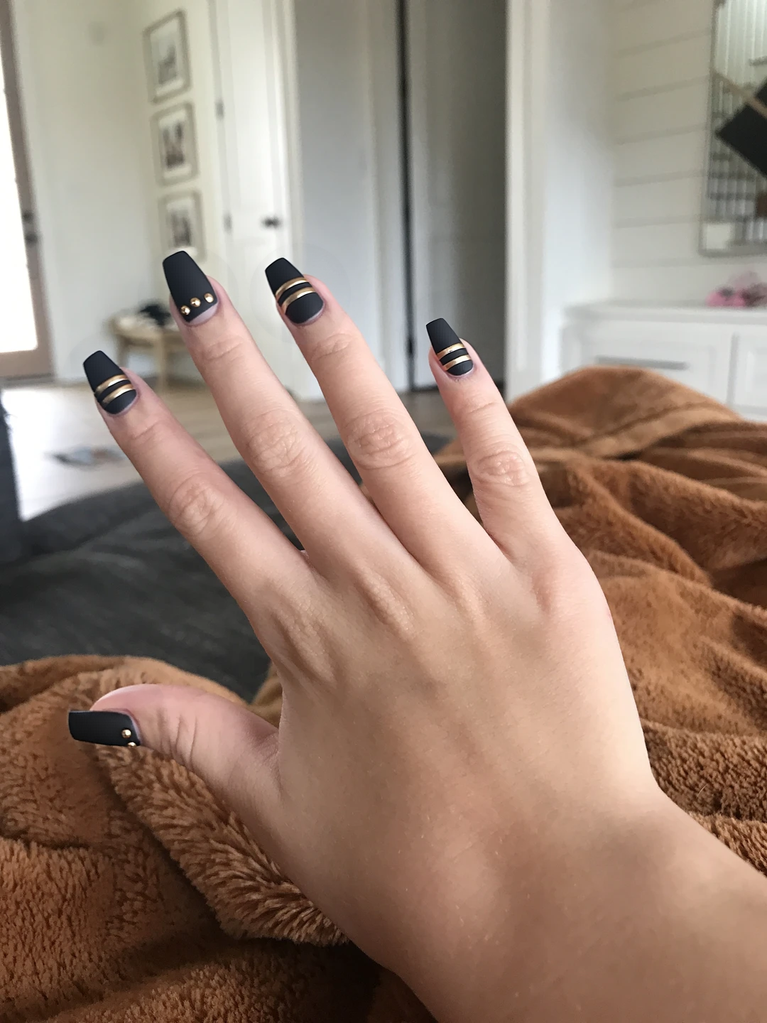A sleek matte black nail design featuring glossy stripes and dots for a high-contrast look. The base of each nail is a deep matte black, providing a sophisticated background. On the ring fingers, three thin glossy stripes run diagonally from the cuticle to the tip, creating an elegant, modern touch. The other nails showcase two glossy dots placed near the cuticle, offering a subtle yet striking accent. The overall finish is matte for the nails with a smooth texture, while the glossy elements add a refined shine, enhancing the sleek aesthetic.
