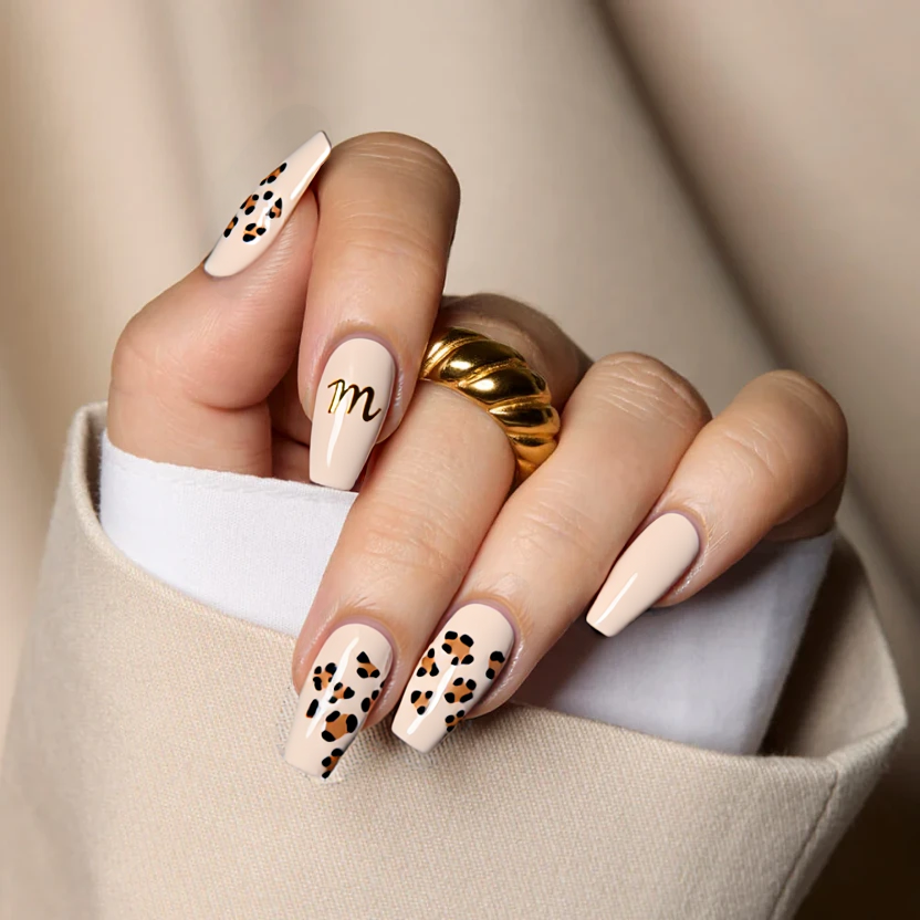 Nails featuring a classic French tip design with a soft, creamy nude base. The tips are adorned with a delicate leopard print pattern, displaying a combination of subtle tan and light dusty brown spots, creating a chic and playful look. The ring finger stands out with a tiny, elegant 'M' delicately hand-painted in a soft gold hue, positioned near the cuticle for a personalized touch. The remaining nails maintain the minimalist leopard print tips, ensuring a cohesive but stylish look. Finished with a glossy topcoat, this design harmonizes perfectly with the medium square shape, offering a trendy yet sophisticated vibe.