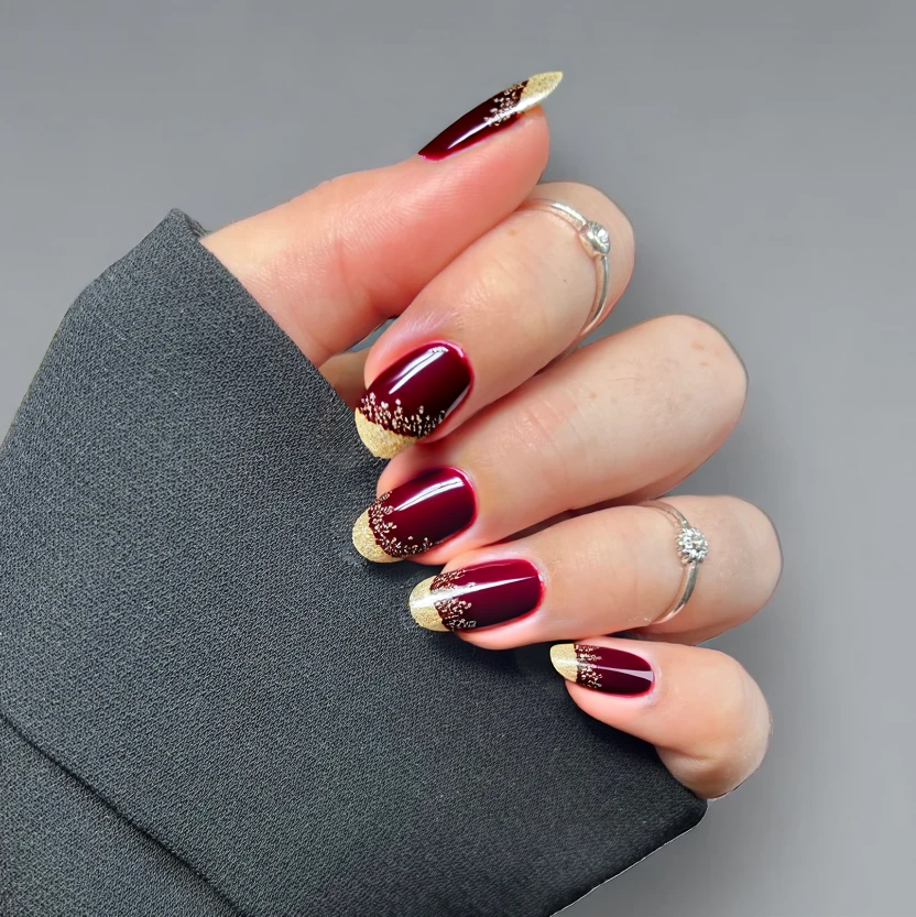 Nails adorned in a rich maroon base color, creating a warm and inviting background for the design. Delicate sparkly accents, featuring fine gold glitter, are subtly scattered across the tips of each nail, adding a touch of elegance without overwhelming the simplicity of the color. The almond shape enhances the gentle curves of the nails, ensuring the overall look remains feminine and refined. A glossy finish completes the design, giving the nails a lustrous and polished appearance.