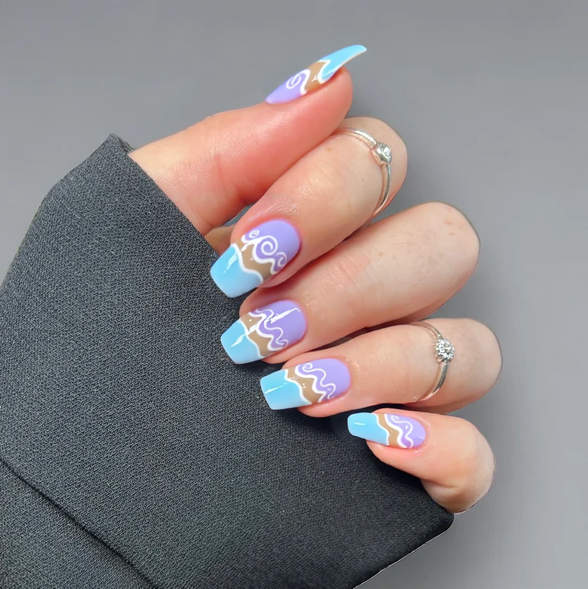Nails painted in a soft lavender base that gently transitions into a light blue at the tips, creating a serene ombre effect reminiscent of ocean waves meeting a sandy beach. On the index and ring fingers, delicate, minimalist white wave patterns swirl gracefully across the surface, evoking a tranquil seaside ambiance. The remaining nails retain the ombre effect without additional designs, ensuring a balanced look. A glossy finish enhances the overall elegance, reflecting the calming essence of a beach getaway, perfectly suited for the short almond shape.