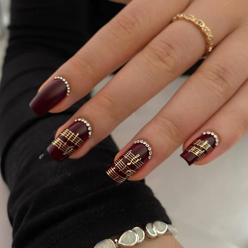 Nails painted in a deep, dark red that captures the essence of elegance and passion, providing a chic backdrop. On the ring fingers, delicate music notes are subtly hand-painted in a soft gold to add a classy touch, limited to just two notes per nail for simplicity. Tiny, clear crystals are placed at the tip of each accent nail to create a hint of sparkle without overwhelming the overall look. The remaining nails are left with the same dark red base, ensuring a cohesive and refined style. A glossy finish enhances the allure of the bold color and details, perfectly tailored to the sharp yet petite nature of short stiletto nails.