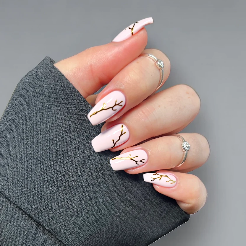 Nails in a soft, pale pink base color that exudes elegance, perfectly suited for short almond-shaped nails. The index and ring fingers feature delicate, minimalistic gold foil accents that create a subtle yet classy design, resembling whispering vines. The other nails remain plain with the same pale pink finish, allowing for a balanced and sophisticated look. A glossy topcoat enhances the shine while maintaining an understated appearance, ideal for an elegant yet simple style.