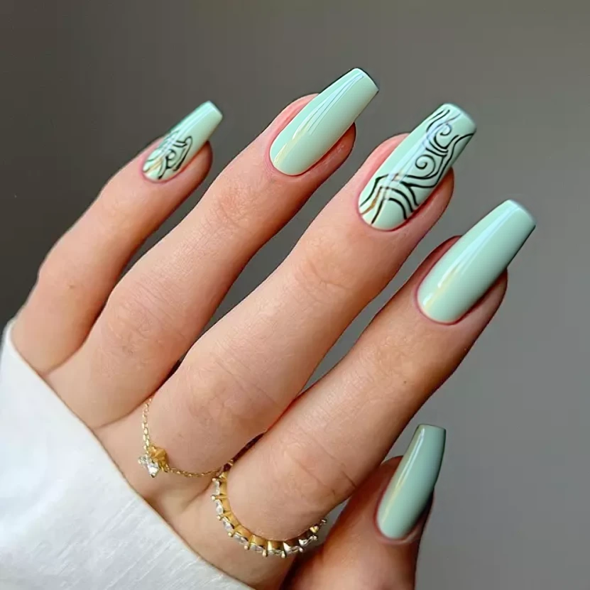 Nails adorned in a soft mint green base color, evoking a serene and fresh aesthetic. On the ring fingers, delicate, abstract swirls in a slightly darker shade of sage green weave gracefully along the length of the nail, creating an elegant, flowing pattern. The remaining nails showcase the mint green hue without any additional textures, maintaining a clean and minimalist look. A glossy finish enhances the overall lightness of the design, perfectly accentuating the dramatic, sharp shape of the medium stiletto nails.