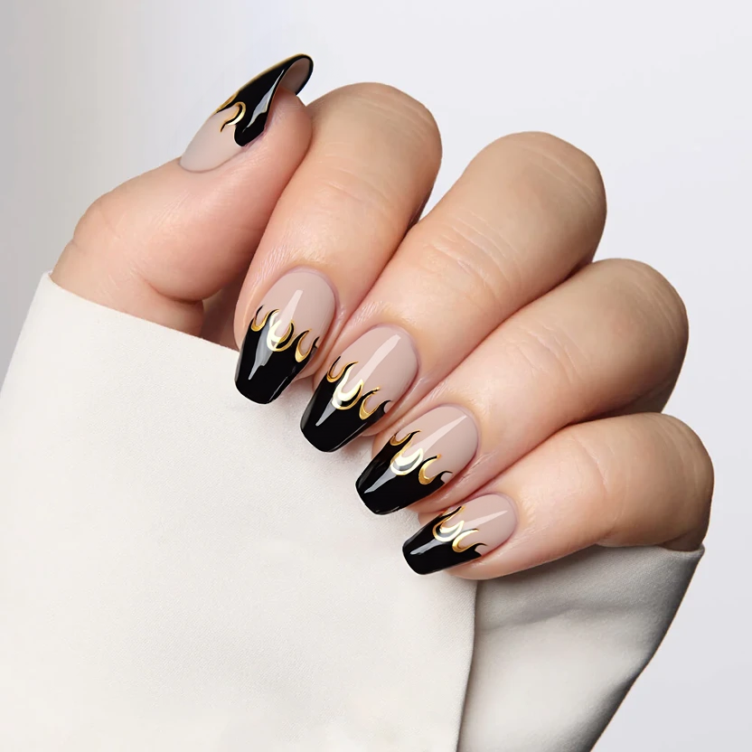 Nails feature a sheer nude base, providing an elegant and understated foundation. The almond-shaped nails are adorned with sharp black French tips that create a striking contrast, emphasizing the gentle curve of the nail's edge. On the ring fingers, delicate accents of small gold foil flames gracefully rise, adding a touch of flamboyance without overwhelming the design. The overall finish is glossy, enhancing the sophisticated look while maintaining a feminine charm that perfectly complements the medium length of the nails.