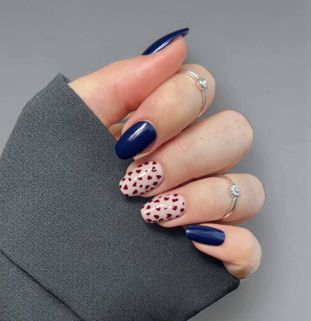 Nails featuring a classic dark blue French tip that elegantly curves along the free edge, complementing the almond shape. The remaining nail surface is adorned with small, delicate dark red hearts scattered modestly across a soft nude base, creating a charming contrast without overwhelming the design. The overall finish is glossy, enhancing the rich colors while maintaining a sophisticated and feminine look, perfect for the understated elegance of short almond nails.