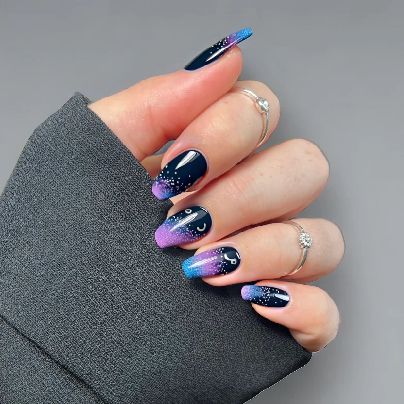 Nails featuring a deep gradient transitioning from dark navy blue at the base to a rich purple near the tips, creating a cosmic night sky effect. The background remains smooth, while the accent nails showcase delicate, minimal glitter sprinkled across the surface to mimic twinkling stars. On one accent nail, a small, elegantly painted crescent moon in silver adds a whimsical touch, while the other designs remain simple and cohesive. The overall glossy finish enhances the celestial theme, perfectly complementing the gentle curves of short almond-shaped nails.