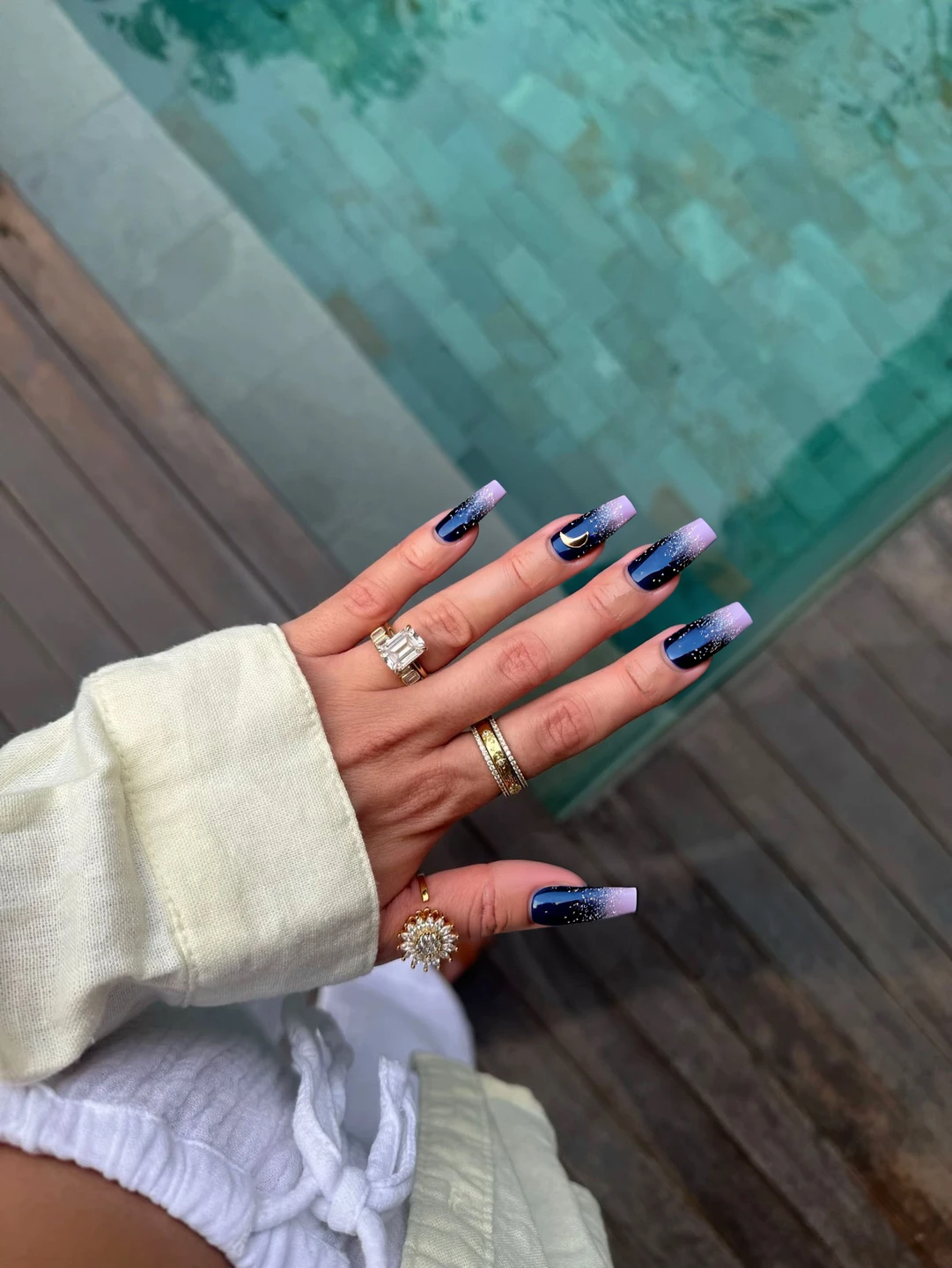 Nails featuring a rich gradient that transitions from deep navy blue at the cuticle to a soft lavender at the tips, creating a cosmic sky effect. On the index and pinky fingers, a scattering of fine silver glitter mimics shimmering stars across the gradient backdrop, adding a celestial sparkle. The middle finger boasts a delicate accent of a hand-painted small silver crescent moon, positioned near the cuticle, enhancing the cosmic theme. The overall design is complemented by a glossy finish that gives the illusion of a night sky, perfectly suited for the elongated elegance of stiletto nails.